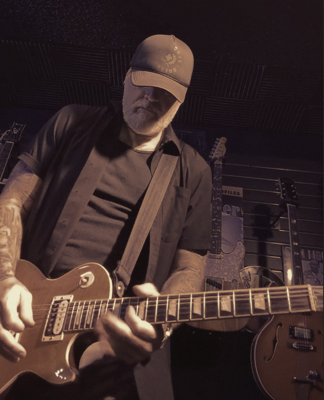 Low-angle close-up of Matthew looking down at the neck of a Les Paul, a wall of hanging guitars behind him.
