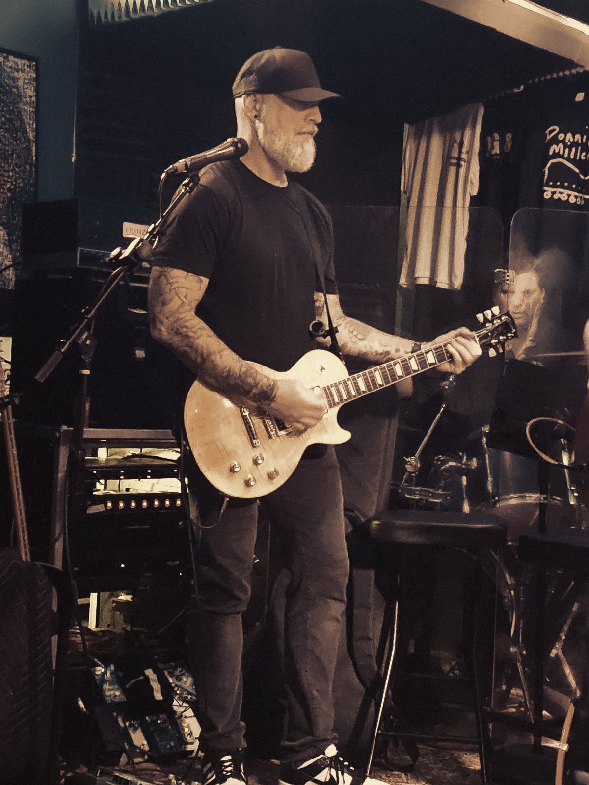 Matthew standing full-length on a small club stage, looking down at the fretboard of a flame-top Les Paul, a mic stand to his left and a pedalboard at his feet.