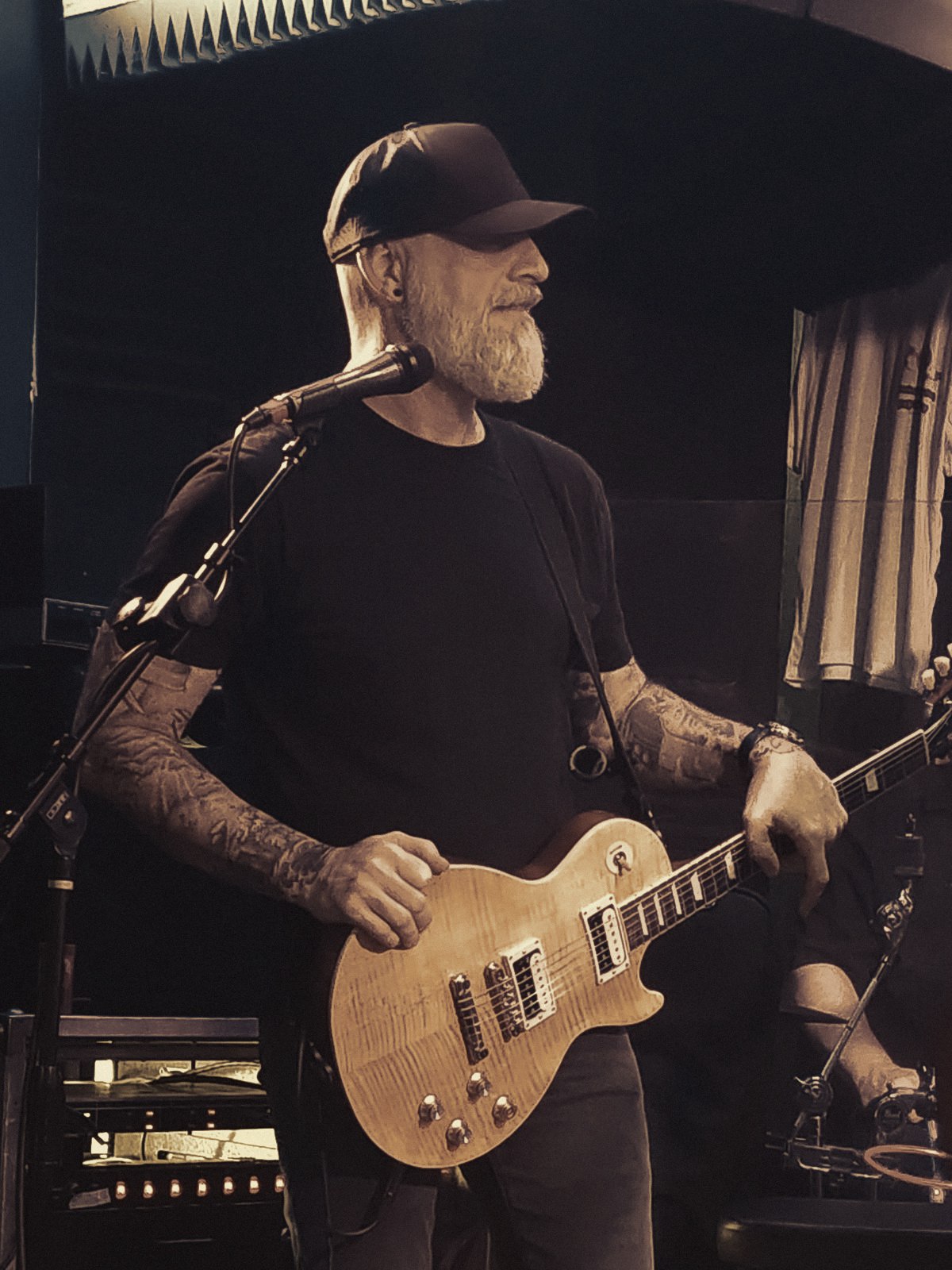 Matthew Sloan Wallace in profile at a microphone, black ballcap and silver beard, holding a flame-top Les Paul under warm stage light.