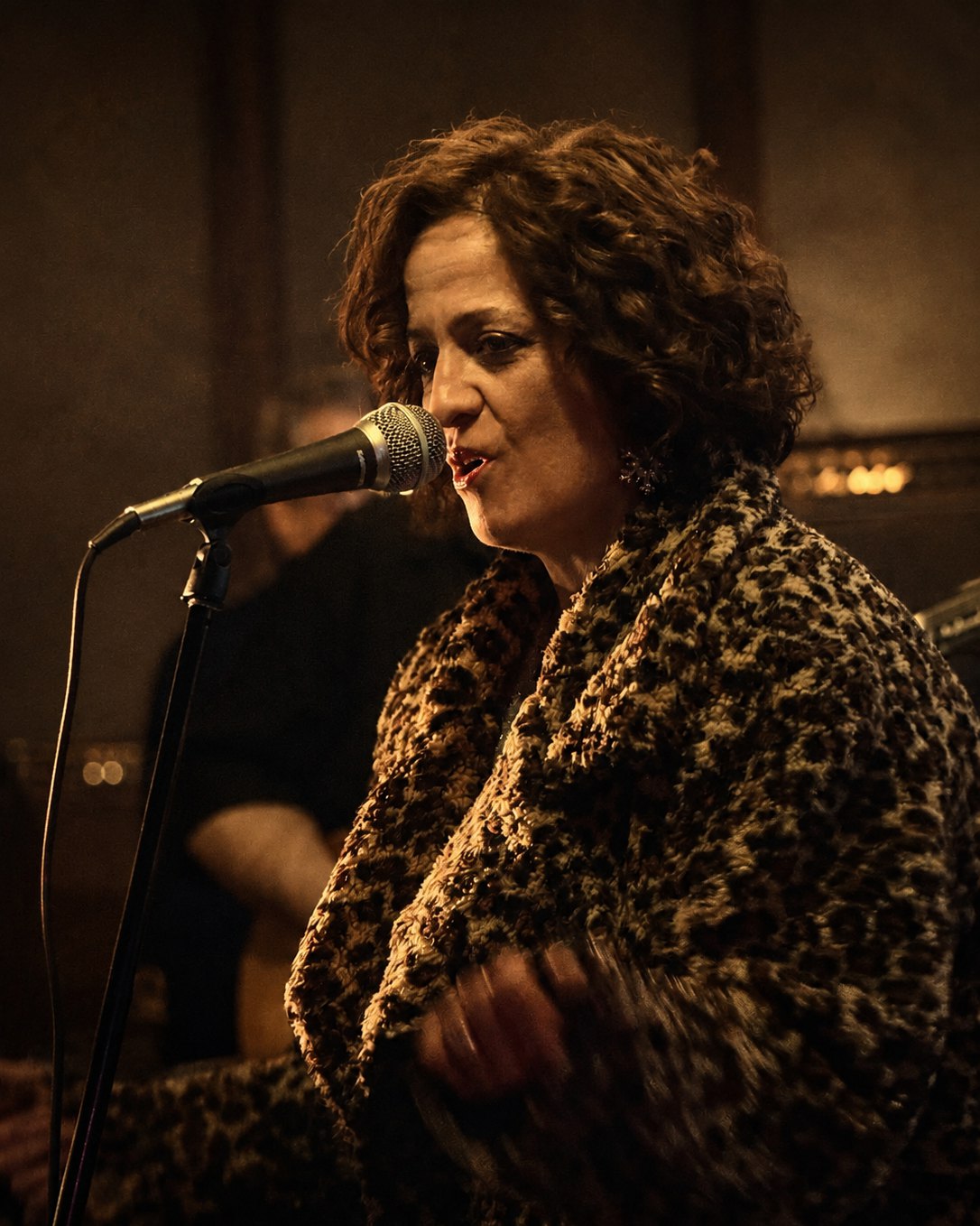 Amanda Chun singing into a handheld microphone with her eyes closed, in a leopard-print coat in a dim wood-panelled room.