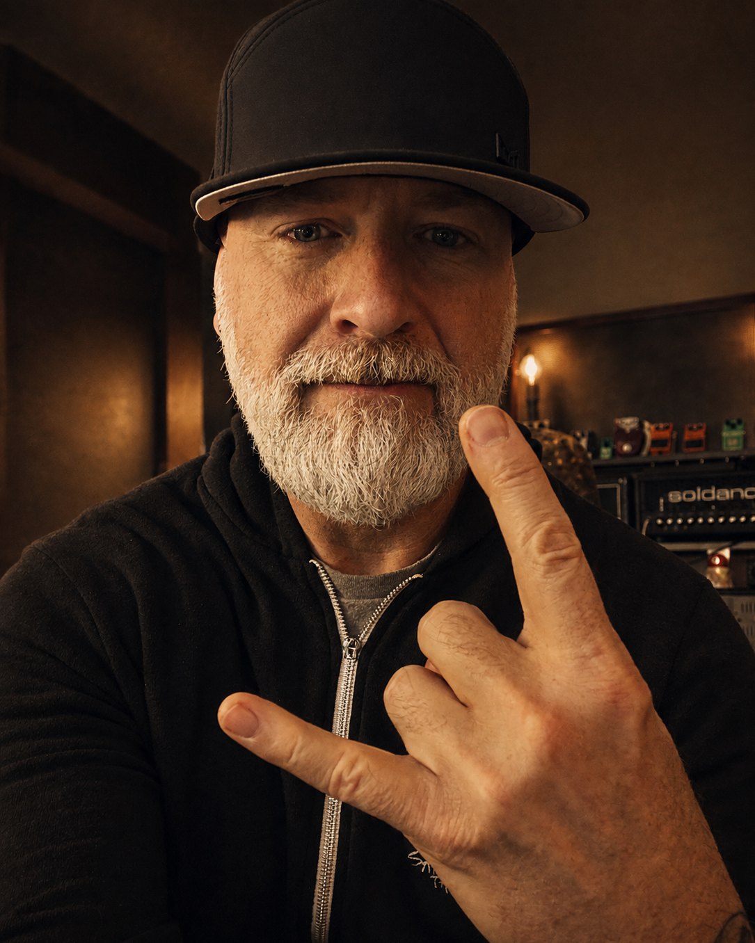Matthew Sloan Wallace in a black cap and zip hoodie looking straight at the camera, a Soldano amp head and a shelf of effects pedals behind him.