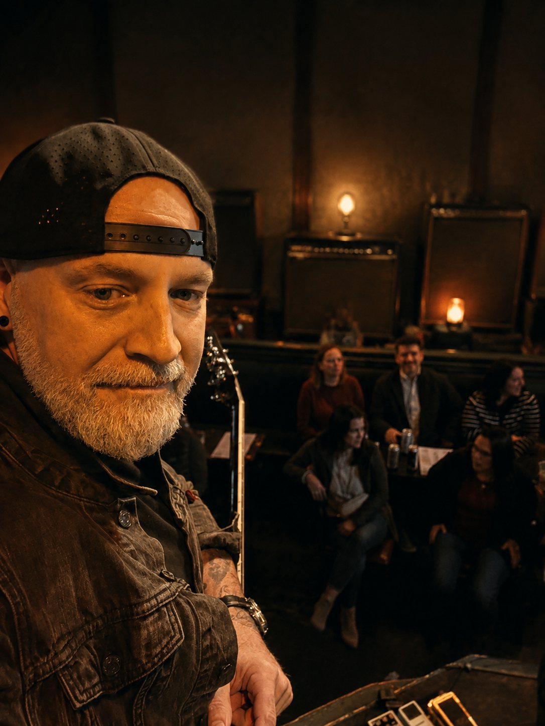 Matthew with his cap on backwards, in a dark denim jacket, looking back over his shoulder from the stage toward a small seated audience in a candle-lit room.