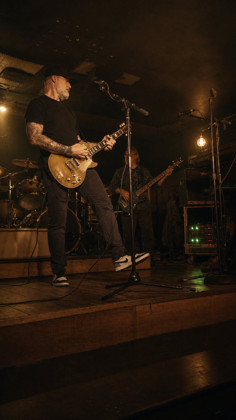 Matthew Sloan Wallace in a black cap playing a goldtop Les Paul at a microphone stand on a plank stage, drums and a bassist behind him.